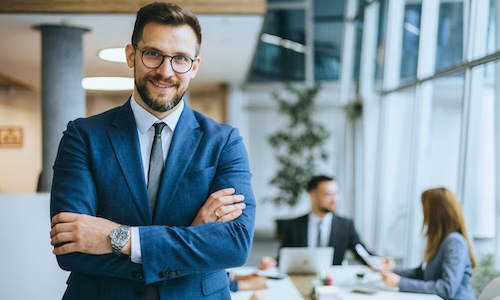 An executive business person exudes confidence in formal businesswear, with a corporate office setting and colleagues engaging in teamwork in the background